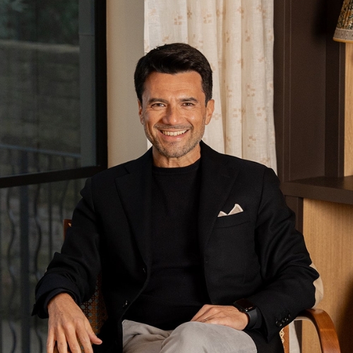 Image of a man in a black shirt and suit jacket sitting in a chair in front of a window