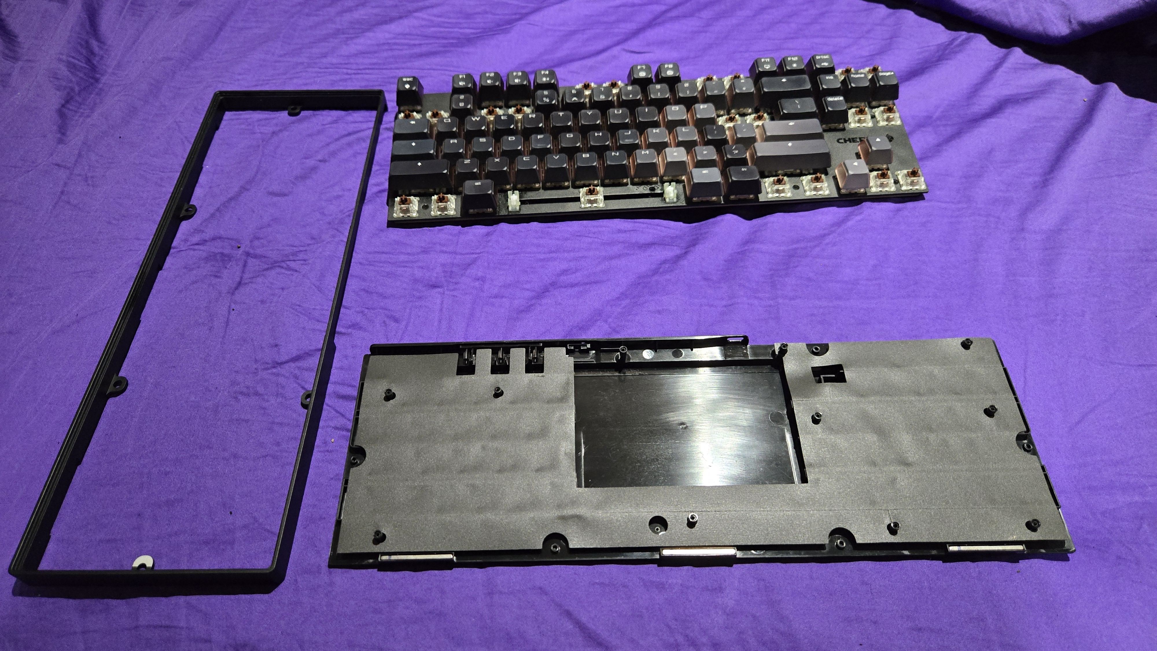 The Cherry MX 8.2 Wireless Xaga in pieces on a purple sheet. We see the bezel ring, the keycaps, and the bottom tray obscured by what appears to be a layer of sound dampening foam.