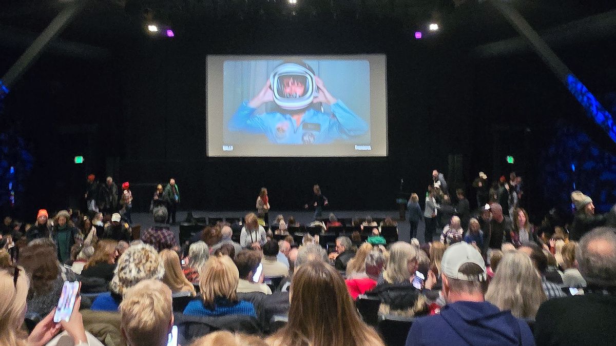 an audience in a theater views a screen with a still image of a female astronaut