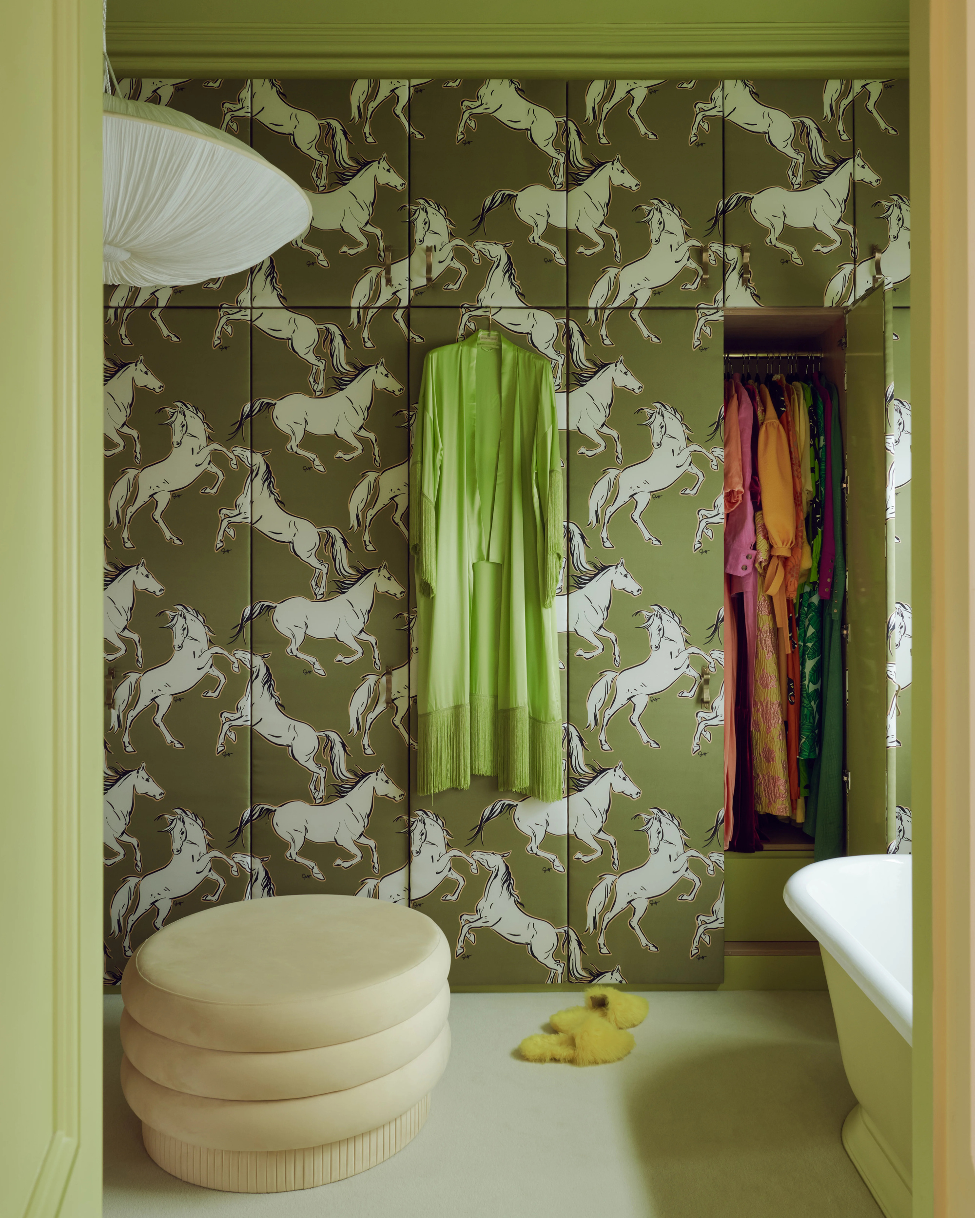 dressing room with green wallpaper on closet doors with horse print, a dress hanging down, an ottoman, fluffy slippers on the floor, a pendant light, and bath tub
