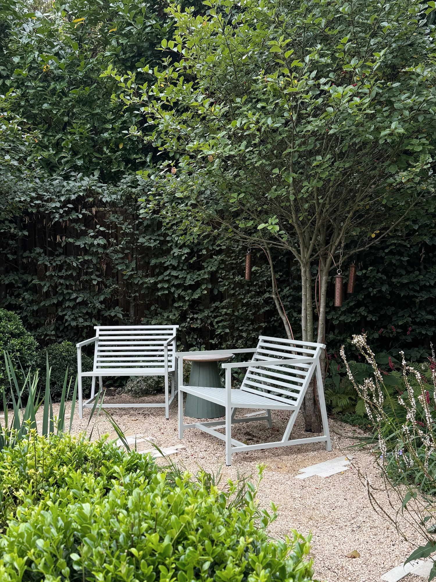 Evergreen garden seating area with slatted metal armchairs, gravel and pavers