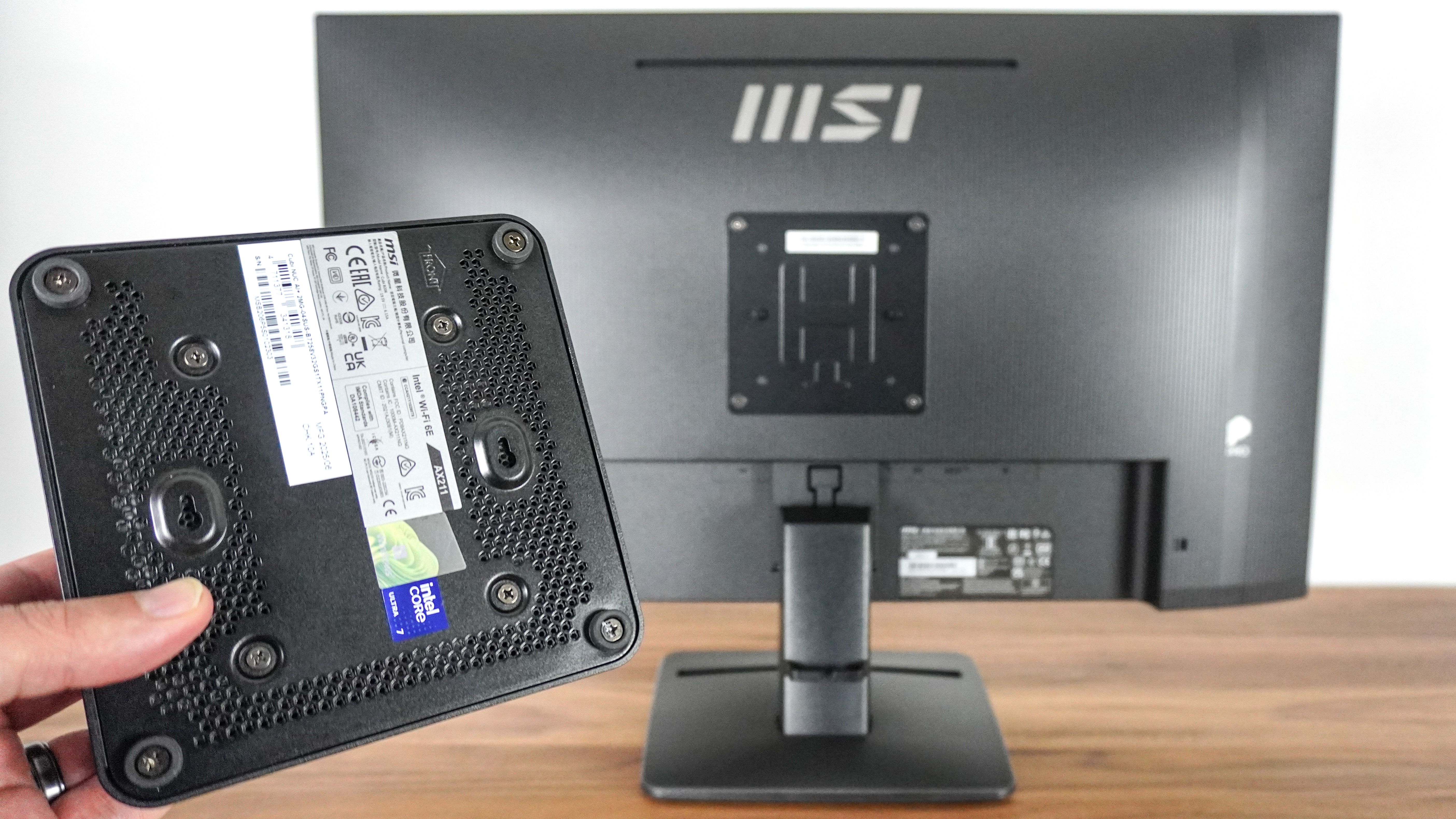 A person holding the MSI Cubi NUC AI+ 2MG mini PC in front of an MSI monitor with a VESA mounting bracket installed on the back of the display
