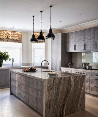 A dark grey marble island in a kitchen with paisley blinds and pendant lights
