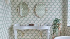 A his and her bathroom sink with two ovel mirrors above each sink. There is pretty green and white wallpaper on all of the walls and a rattan stand with a trailing plant sitting on top.
