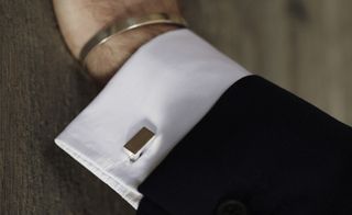 The wrist of a man with a suit on wearing square silver cuff links and a silver bracelet.