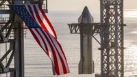 A giant American flag flutters in front of a silver SpaceX Starship rocket as it is hoisted atop its booster.