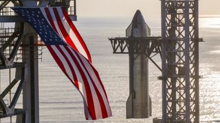 A giant American flag flutters in front of a silver SpaceX Starship rocket as it is hoisted atop its booster.