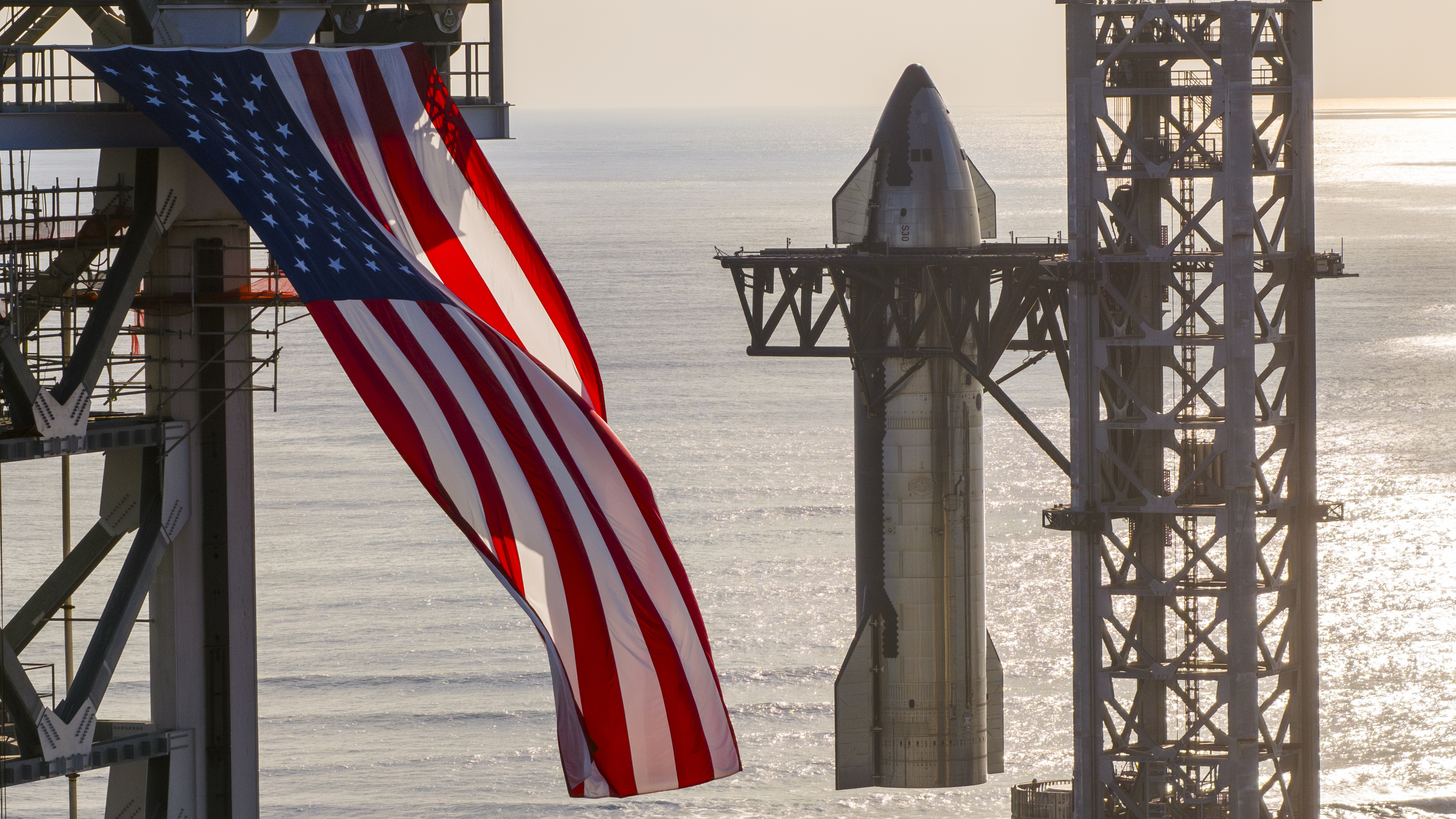 A giant American flag flutters in front of a silver SpaceX Starship rocket as it is hoisted atop its booster.