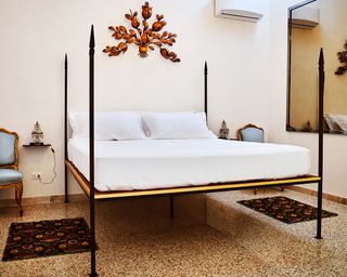 A hotel room with a four post bed, blue chairs, wall mounted side tables, patterned rugs, a wall decoration and a wall mirror.
