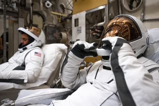 Two astronauts, one in the foreground, the other in the background, both facing left, lean back in white and black bucket sets, wearing white spacesuits.