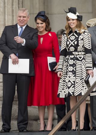 Princess Beatrice wearing a black and white dress and hat standing to the right of Princess Eugenie wearing a red dress and navy hat who is laughing next to Prince Andrew on a staircase