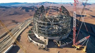 At the centre of this image is the criss-cross steel skeleton structure of the ELT dome, sat on top of a circular concrete base. Tall, spindly cranes are dotted around the circumference of the dome, leaning in all different directions. Past the dome, the brown expanse of the Atacama Desert unfolds into the distance. The desert is a mixture of flat plains and sharp mountain peaks.