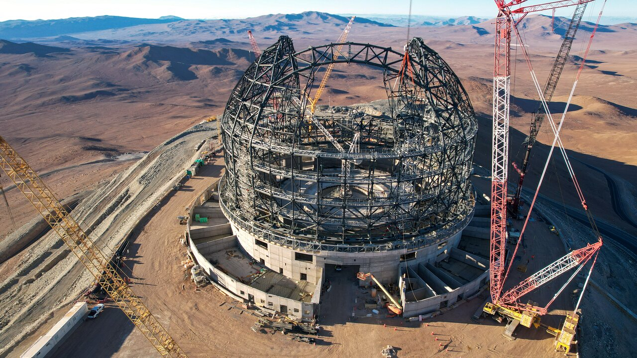 In the center of this image is the cross-shaped steel framework structure of the ELT dome, resting on a circular concrete foundation. Tall, spindly cranes are dotted around the dome, leaning in different directions. Once you pass the dome, the brown Atacama Desert stretches into the distance. Deserts are a mixture of flat plains and sharp mountain peaks.