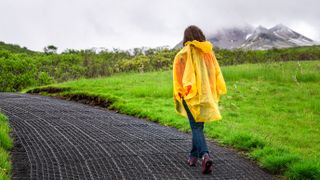 woman in yellow poncho walking hiking on wet trail path hiking road and cloudy sky with mountains