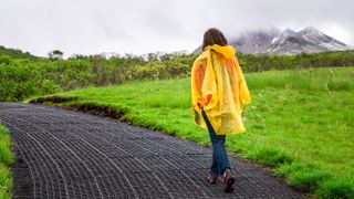 穿着黄色雨披的女人在潮湿的小径上徒步，云雾缭绕的天空中有山