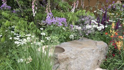 A border blooming with purple, white, and orange flowers behind a stone water feature