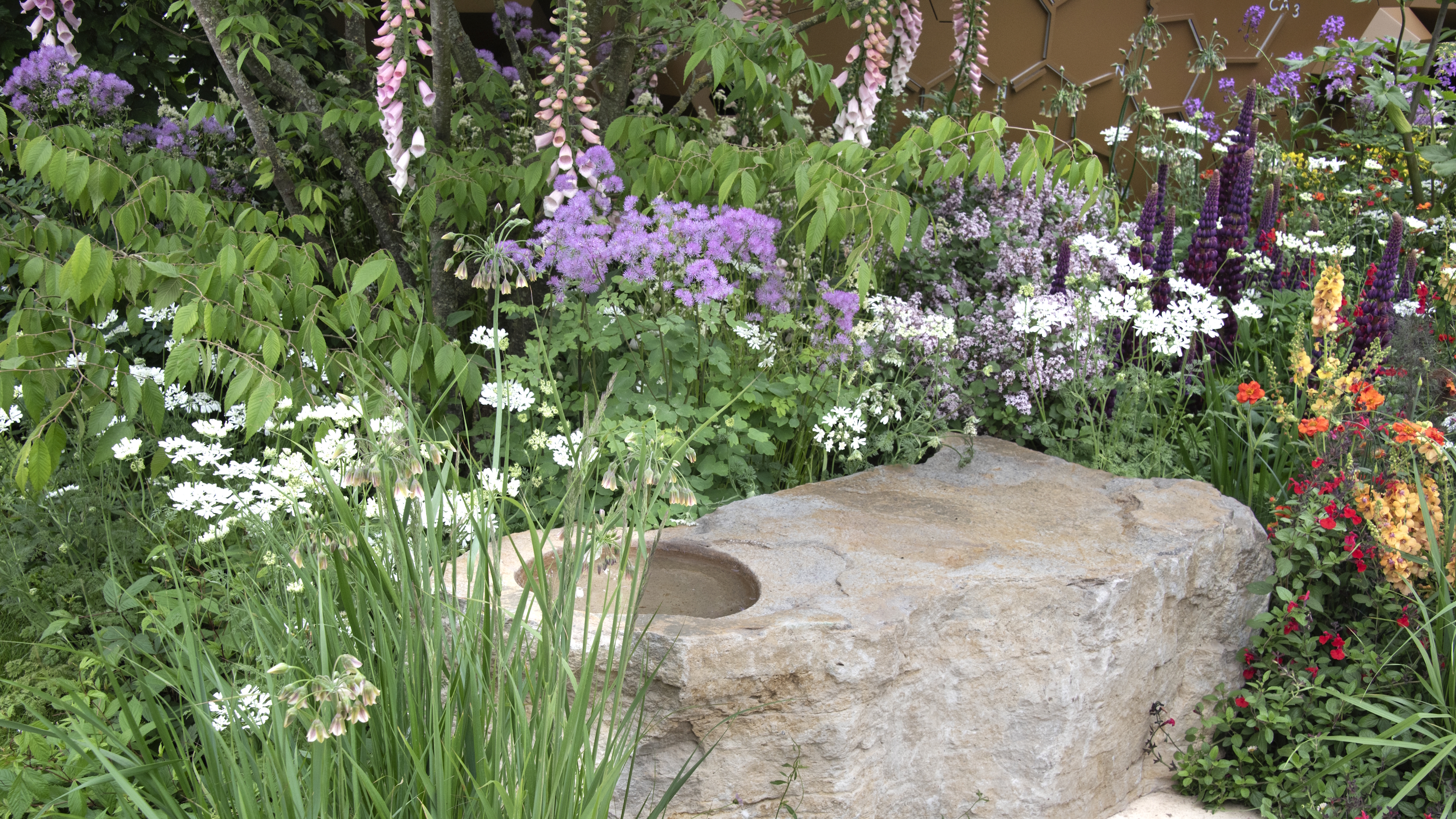 A border blooming with purple, white, and orange flowers behind a stone water feature