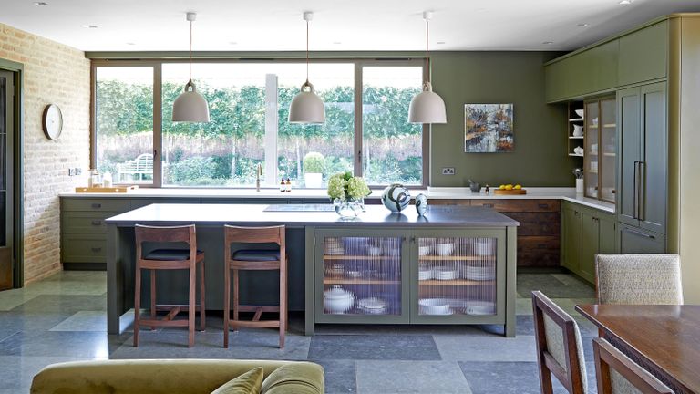Kitchen island storage ideas. Large kitchen with central island, exposed brick features, dark gray tiled flooring, green painted walls, island with storage cabinets with reeded glass doors, three rounded white pendants hanging over island, two dark wood bar stools, dark wood dining table in corner