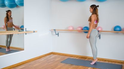 A woman stands on her tip toes on an exercise mat in a dance studio, her right hand on a wooden bar. She is facing a large mirror and the floor is laid with polished wood. 