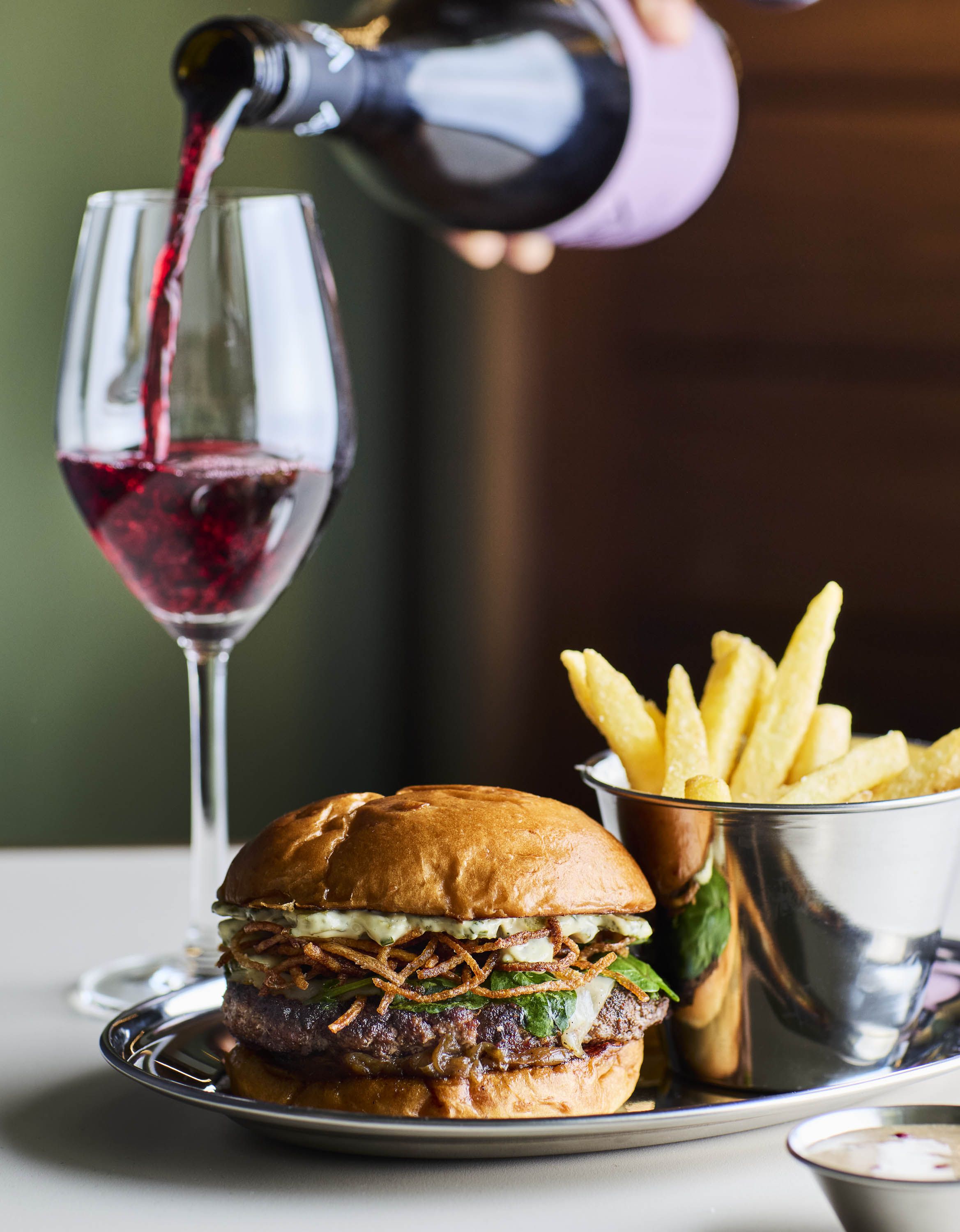 A burger joint's neon-lit sign, a man eating fries and a burger inside it, and a close-up of a chrome platter filled with a large burger and a portion of fries, paired with a glass of wine that's just been poured for the diner.
