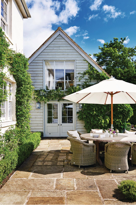 White outdoor umbrella shading a round outdoor dining table and chairs, which sit on a charming stone patio with crawling vines