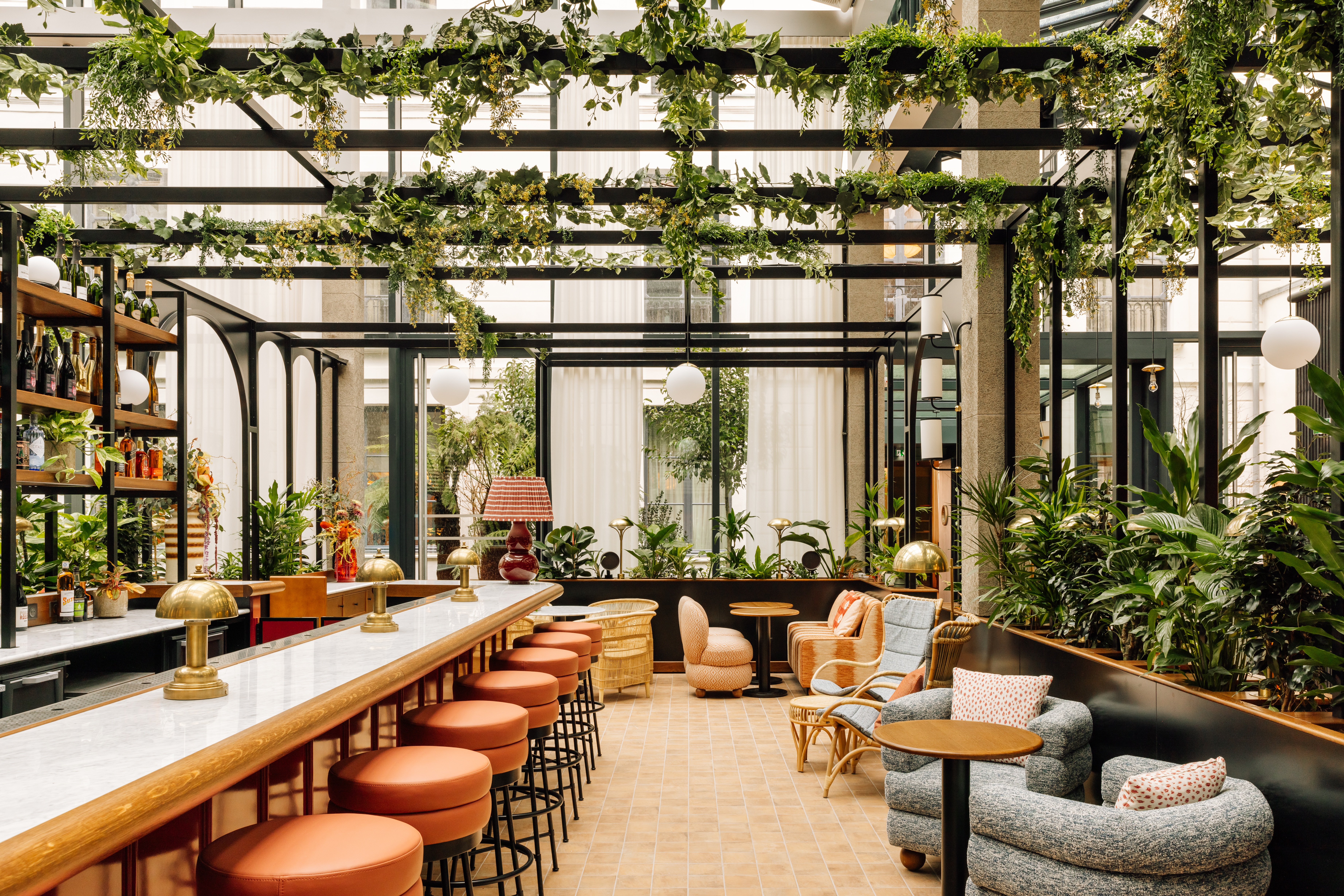 An atrium-style restaurant and bar features floor-to-ceiling windows and wrought iron architectural structures adorned with trailing plants and Art Deco furniture.
