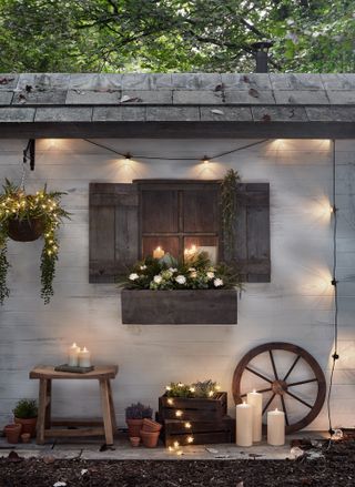 Wooden window box lit up with outdoor lighting surrounded by candles, a stool and hanging planter