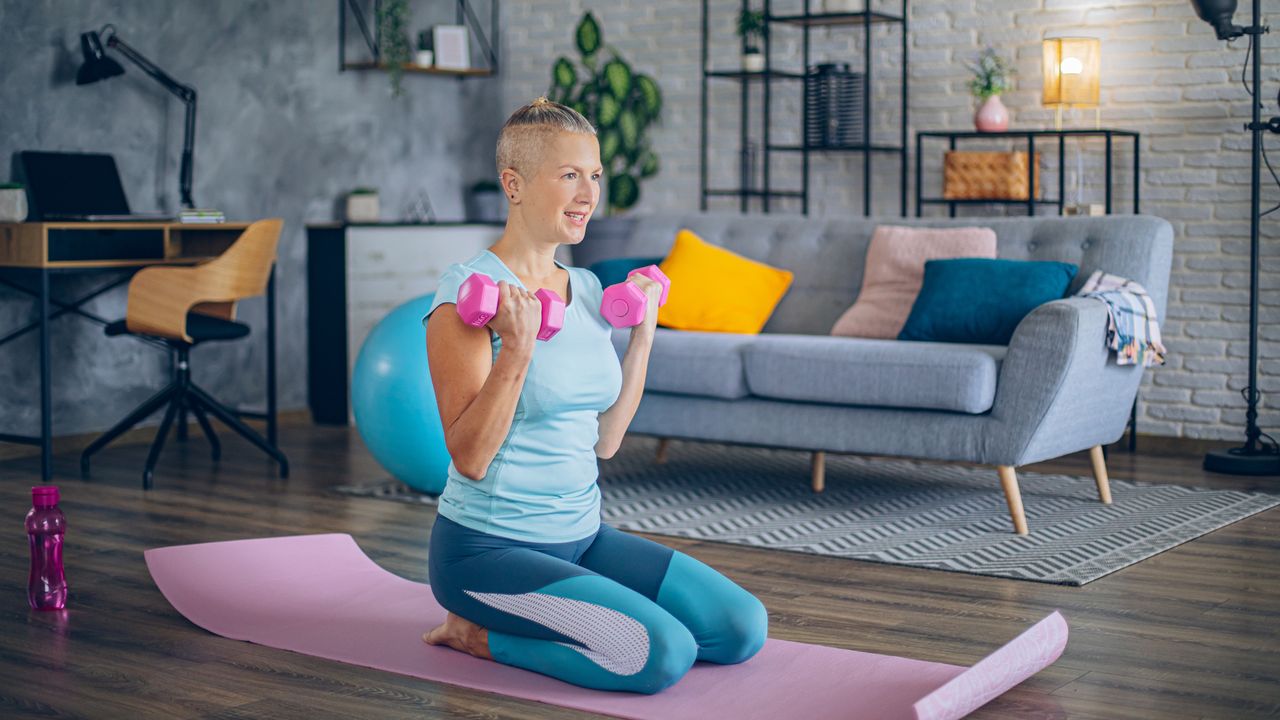 Woman kneels on exercise mat in living room holding pink dumbbells by her shoulders