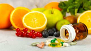 an open bottle of colorful supplements spilled onto a table next to oranges, apples, grapes and blueberries