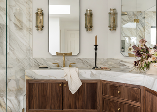 A beige bathroom with wooden cabinets, a marble counter top and luxe wall sconces