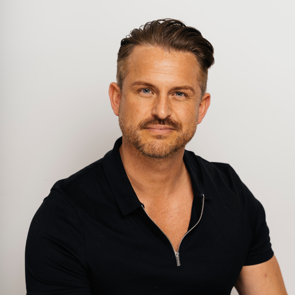 male with short dark hair swept back, facial hair and wearing short black sleeved top with zip