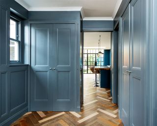 Hallway with painted blue walls and built in cabinets, herringbone patterned dark wood flooring, view looking through hallway to kitchen with kitchen island