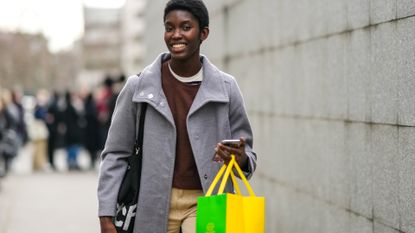 A model wears a white and navy blue striped print pattern t-shirt, a dark brown sweater, a pale gray long buttoned coat, beige large suit pants, a yellow and green shopping bag, outside Chloe, during Paris Fashion Week - Womenswear F/W 2022-2023, on March 03, 2022 in Paris, France