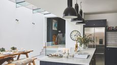 Dark blue kitchen with white walls and a kitchen island with a marble countertop. Three black pendant lights hang above the island and a wooden bench is at the side of the space
