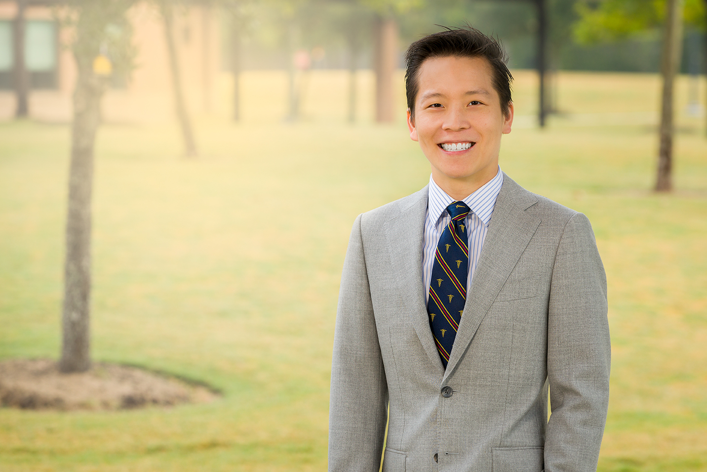 An image of a smiling Chinese doctor in a grey suit outside in the sunshine