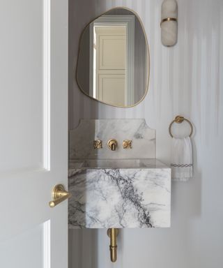 Door open to powder room with neutral veined sink, brass faucets, striped neutral wallpaper and mirror above