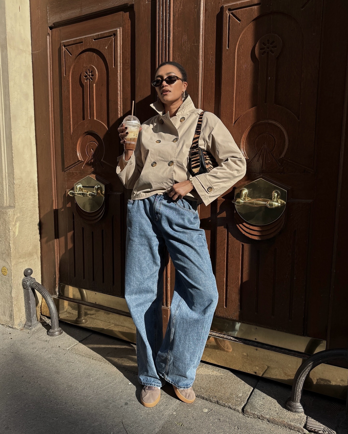 French style influencer Lena Farl wearing oval sunglasses a cropped funnel-neck trench coat, tiger print shoulder bag, baggy jeans, and tan Adidas sneakers