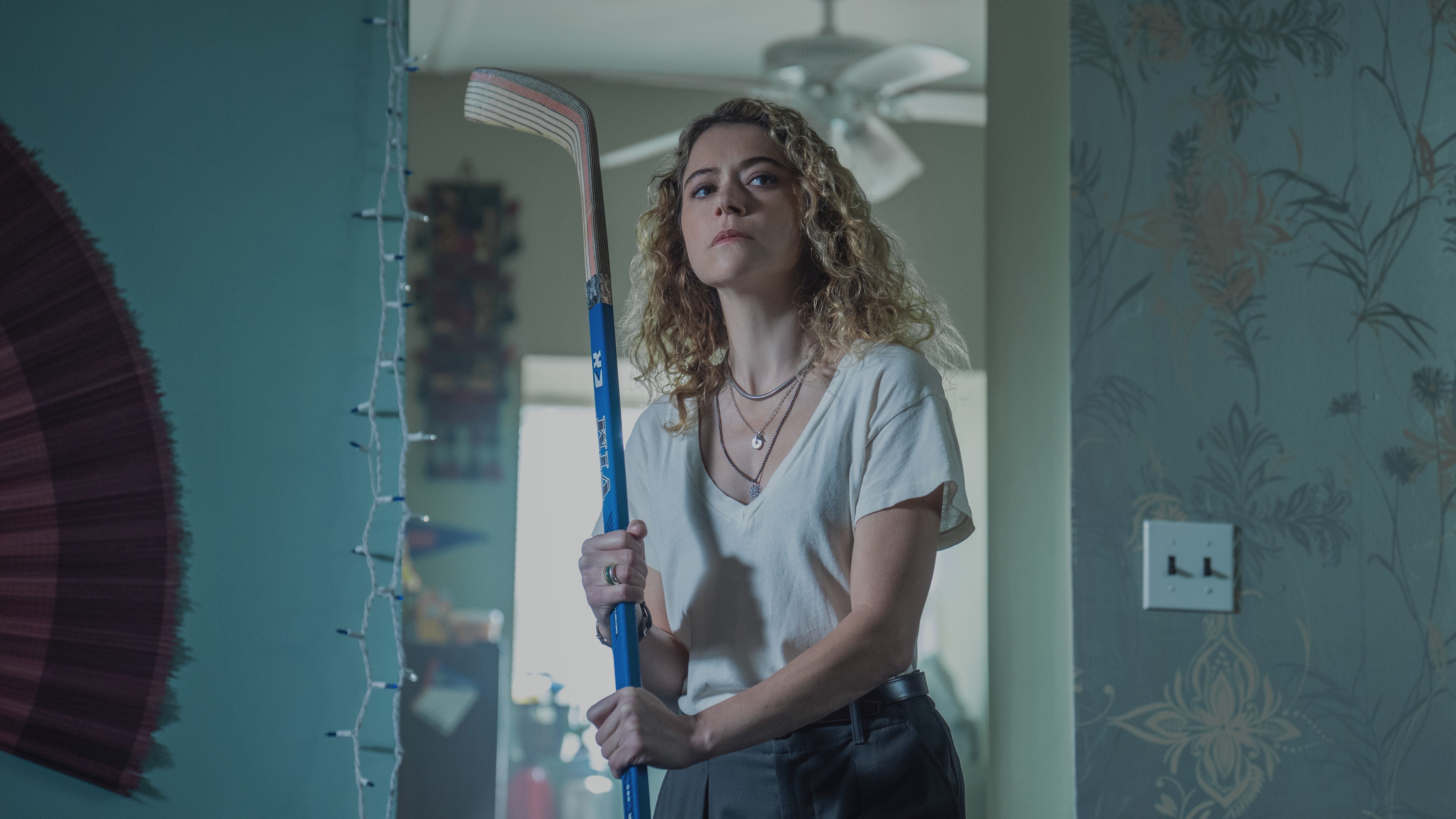 A woman (Tatiana Maslany) brandishes a hockey stick while standing in the doorway of a home, with a string of Christmas lights hung to her right, in the Apple TV series 'Maximum Pleasure Guaranteed.'
