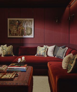 cozy living room with burgundy walls, russet velvet sectional, footstool, fringed couch pillows, artwork, wall lights, books