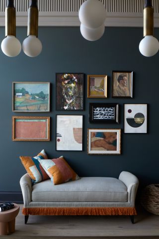 Grey and rust fringed chaise against a dark blue wall filled with gallery wall-style art