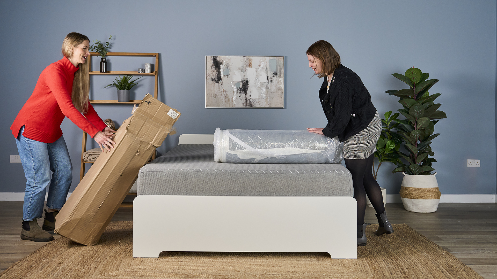 Two testers open a shipping box on a bed to reveal a compressed, rolled-up Helix Dual Comfort mattress topper inside.