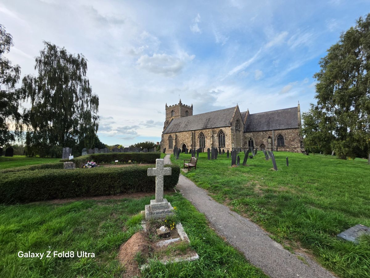 Wide-angle view of a historic stone church and graveyard surrounded by green grass and trees.