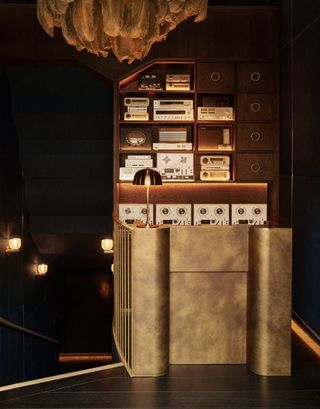 A brass-drenched cocktail bar with a vintage audiophile sound system features Art Deco sconces, mushroom-shaped table lamps, and a sculptural pendant light overlooking a staircase.