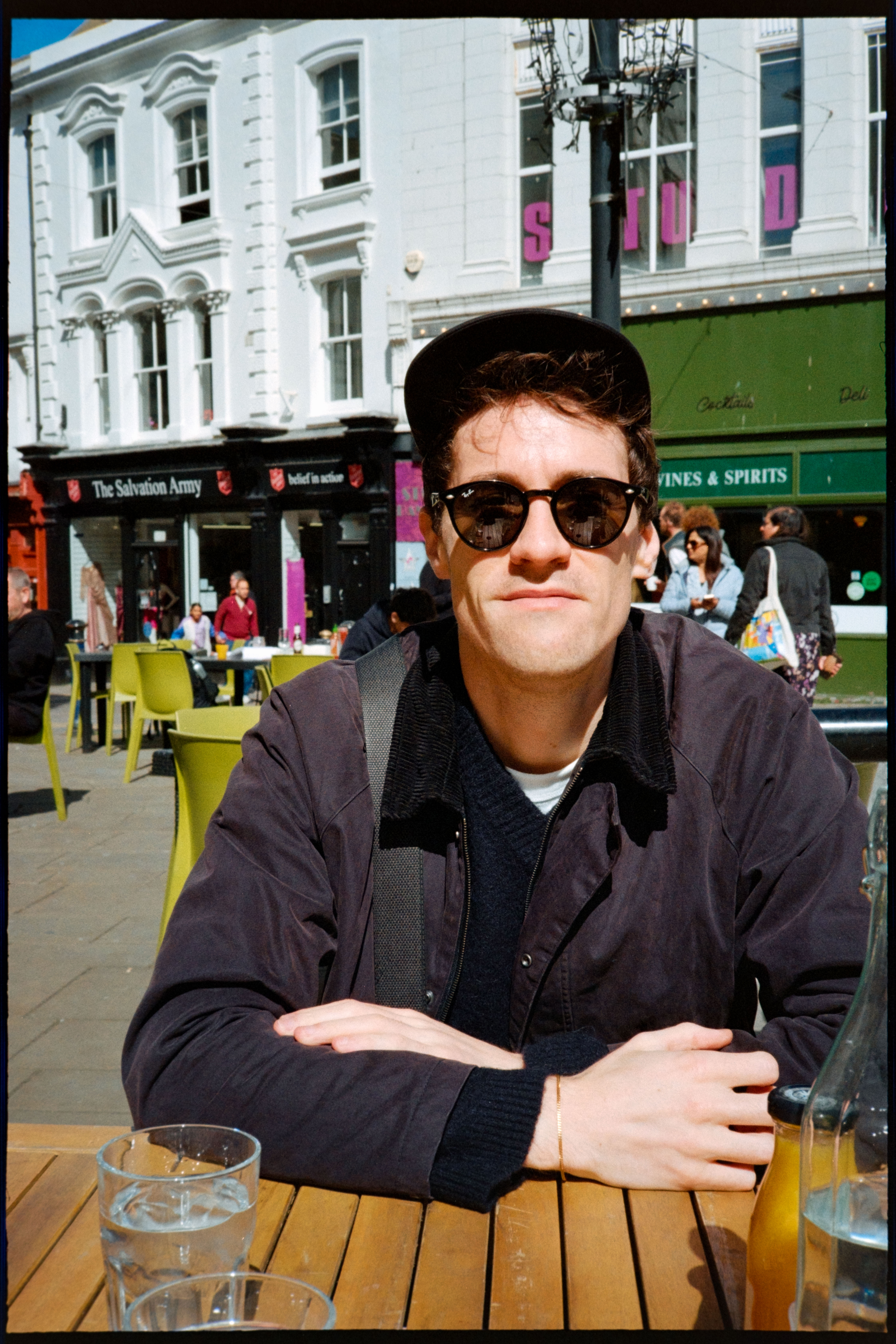 A man wearing sunglasses and a cap sitting at an outdoor caf&amp;eacute; table on a sunny street.
