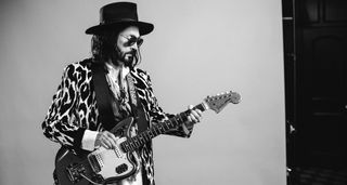 A portrait of Mike Campbell wearing a wide-brimmed hat and dark sunglasses, as the former Heartbreaker posses with a guitar.