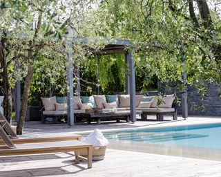 A shaded pergola with plants growing over it above a sofa next to a pool area with sun loungers