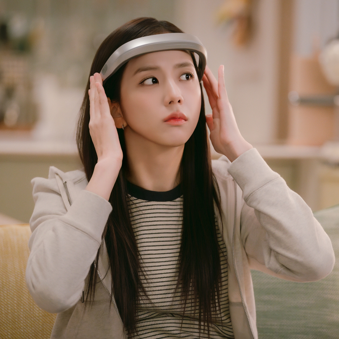 A woman (Jisoo as Seo Mi-rae) holds a headband-style device to her forehead as she sits on a couch in a living room, in the Netflix K-drama 'Boyfriend on Demand.'