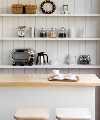 An image of cream shelving unit with crockery and appliances on it, with a kitchen island and stalls in the foreground