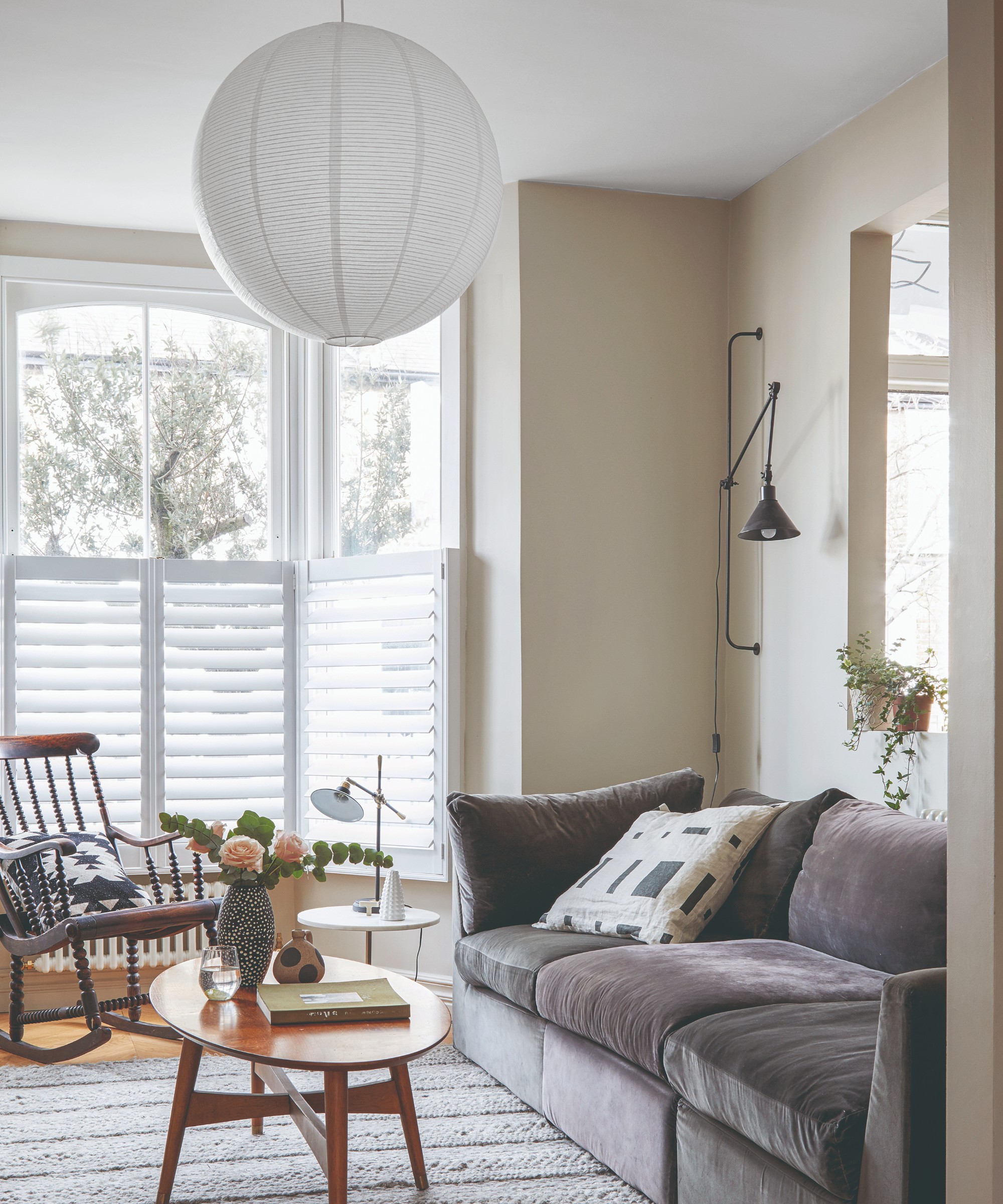 A bright living room with a grey velvet sofa, an adjustable wall light, a table lamp on a side table and a large paper lantern ceiling light