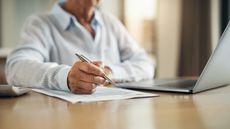 An older man with a pen in his hand works on paperwork at a table with a laptop.